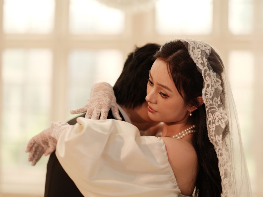 A bride in an elegant lace veil embraces her groom in a romantic indoor wedding setting.