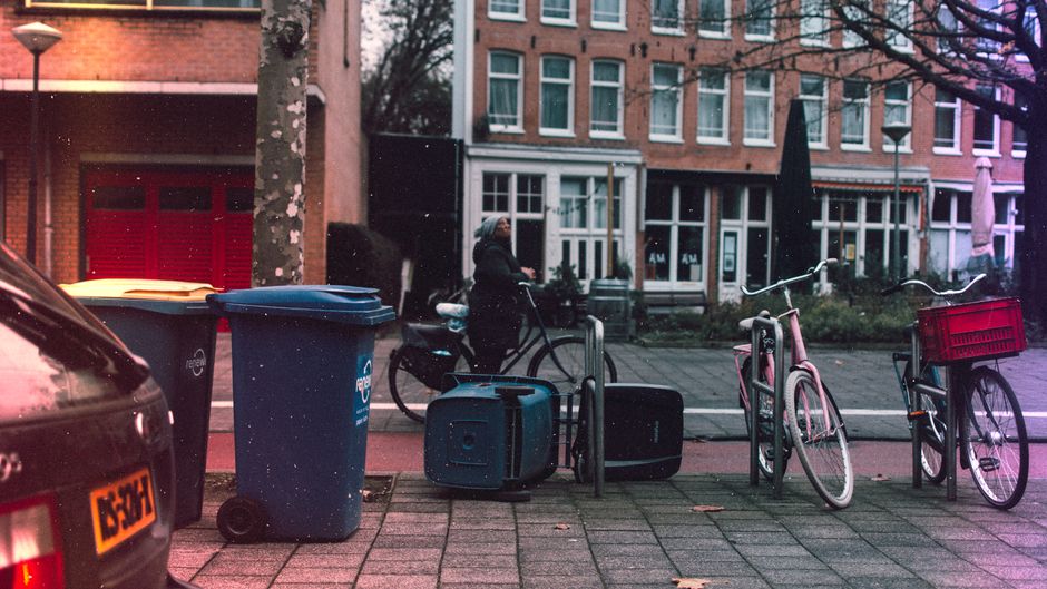 Urban winter street scene with bicycles, bins, and snowflakes falling.