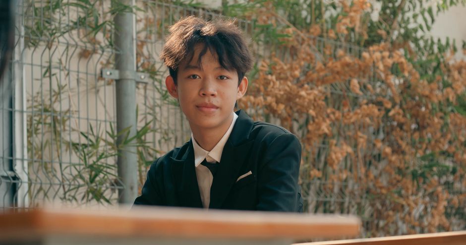 A young adult in a black suit sitting outdoors by a fence, exuding confidence.