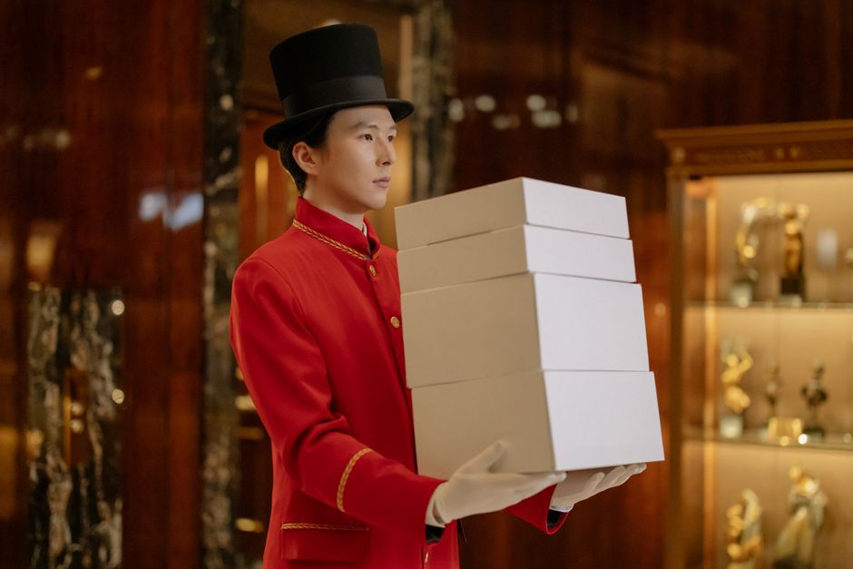 Asian hotel porter in red uniform indoors carrying multiple boxes with a professional look.