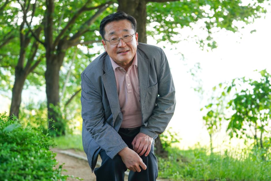 An elderly man wearing a jacket leans forward in a green park setting during summer.