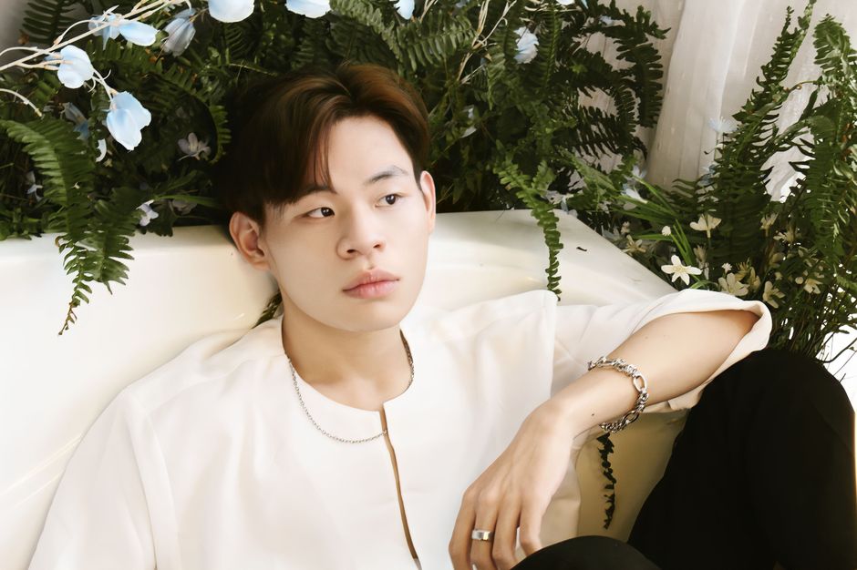 Asian man sitting elegantly on a sofa surrounded by lush greenery. Wearing jewelry.