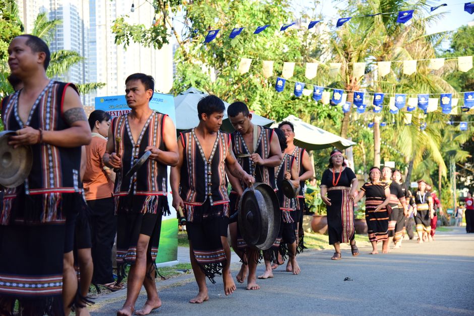 Men and women in traditional attire performing with gongs during a cultural parade outdoors.